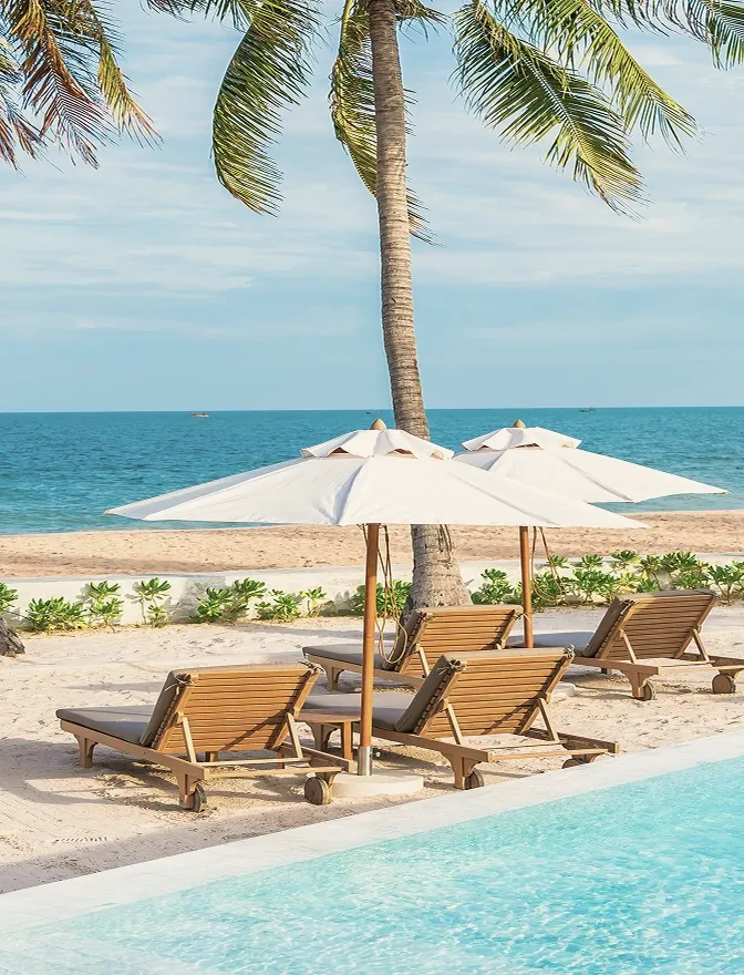 Sun loungers and white parasols on the sand beside the pool, palms above them and the sea beyond