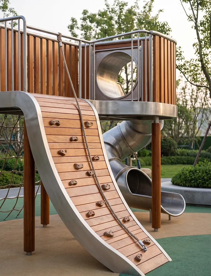 A children's play structure in the grounds: a timber deck, a climbing ramp and a metal slide among the planting